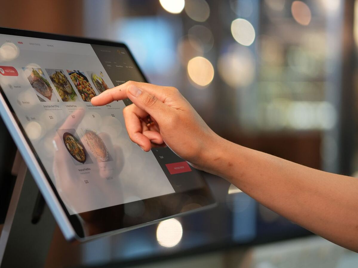 Tangan staf menekan layar kasir yang menampilkan daftar menu bergambar di meja kasir restoran